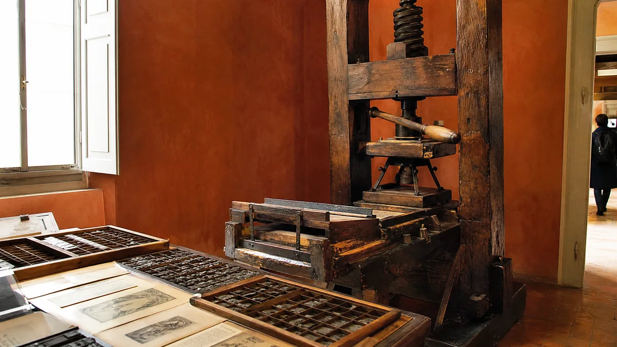 An antique wooden hand printing press beside trays of metal type and old printed pages in a room with terracotta walls