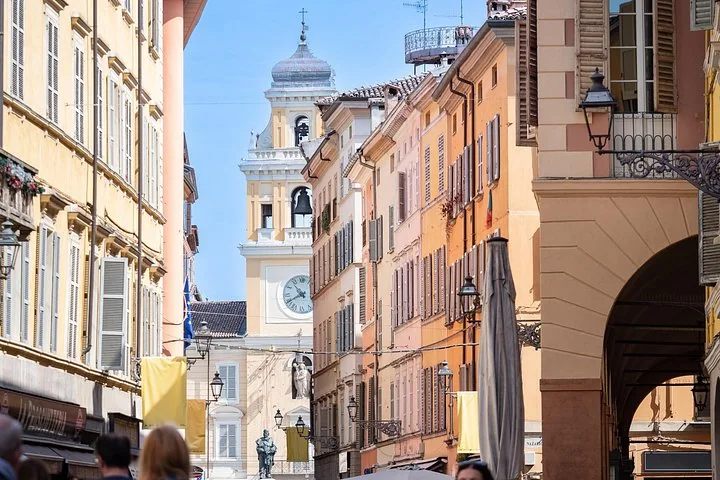 A narrow old-town street of ochre and pink palazzi leading to a clock tower under a blue sky