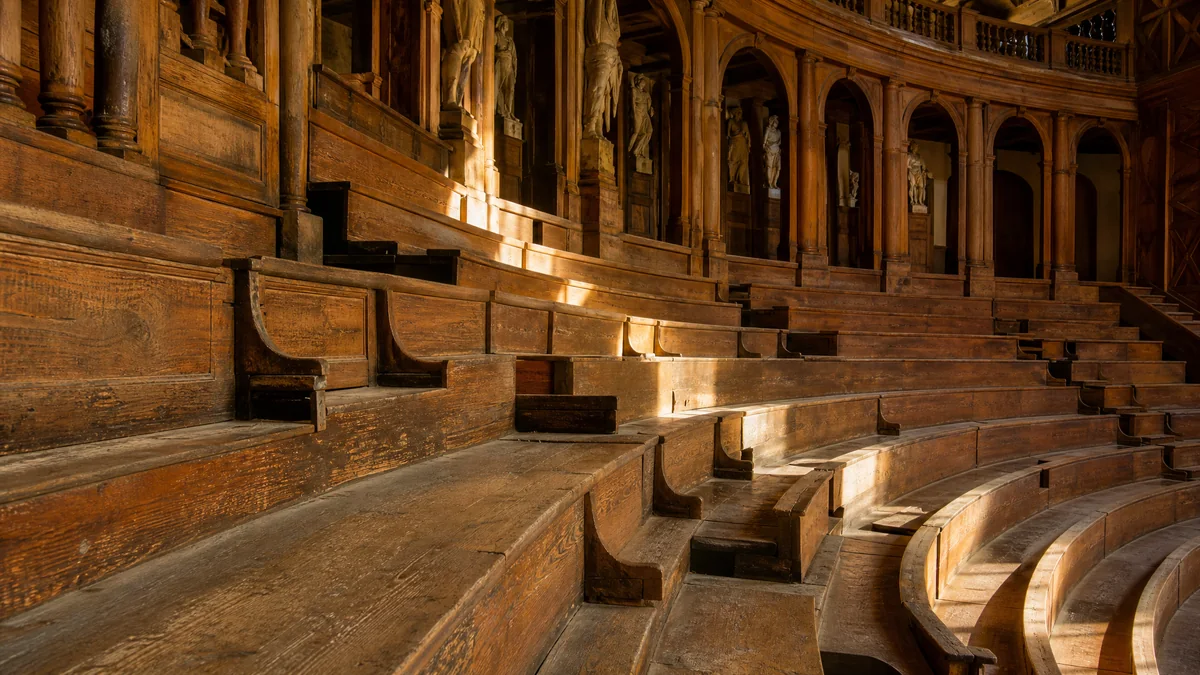 Le ripide gradinate lignee di un teatro di corte rinascimentale sotto i loggiati, in una luce calda